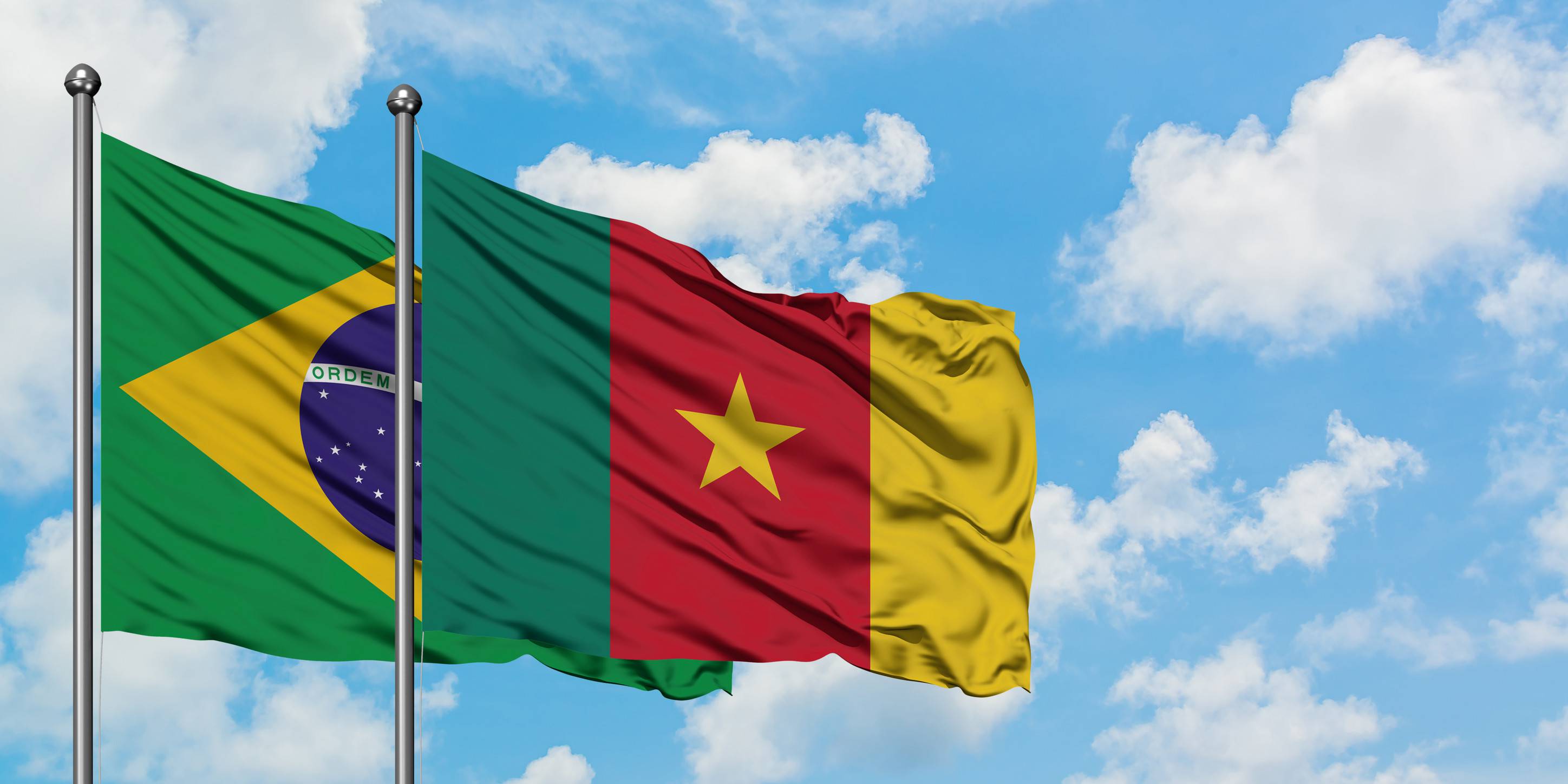 Brazil And Cameroon Flag Waving In The Wind Against White Cloudy Blue Sky Together